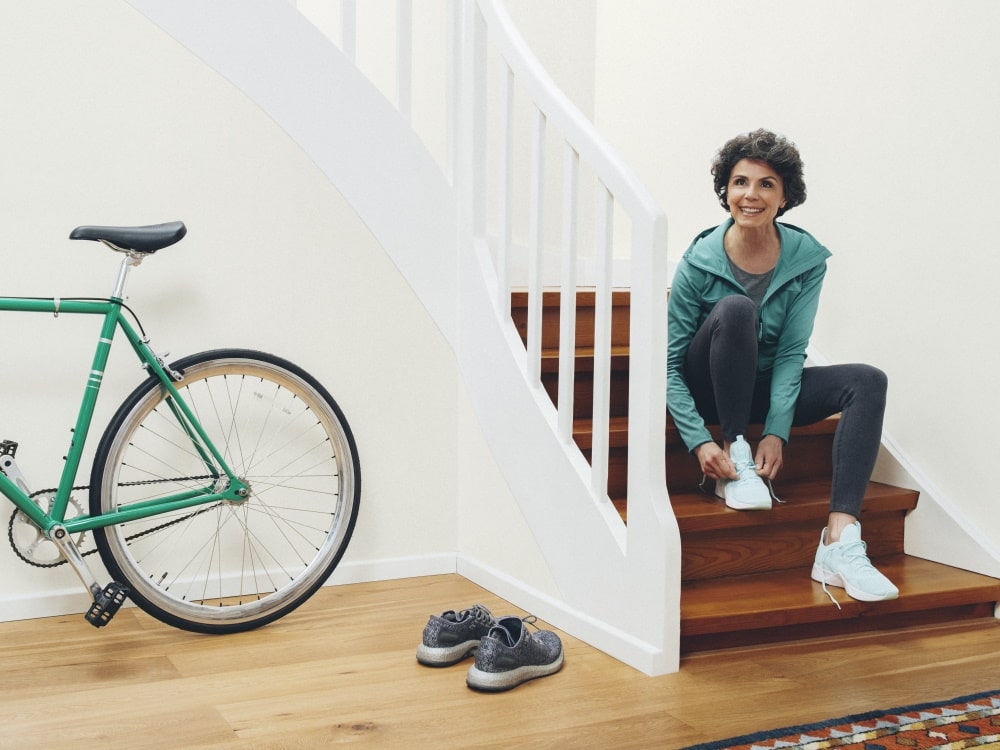 On the right a woman sits on the steps and ties her shoes, on the left there is a bicycle and another pair of shoes